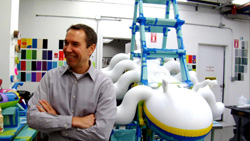 A man with arms crossed stands in a workshop next to a large, white, curved sculpture and a blue ladder structure.