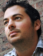 A man with dark wavy hair and light stubble looking up toward the side.
