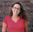 A smiling woman with brown hair and glasses wearing a red v-neck shirt in front of a brick wall.