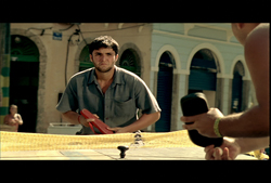 A man in a grey shirt holds a red object while looking forward in a sunlit street.