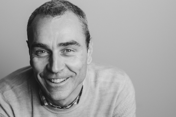 A smiling middle-aged man with short hair wearing a sweater and a collared shirt.