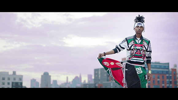 A man in patterned streetwear holds a red and green flag against a city skyline under a cloudy sky.