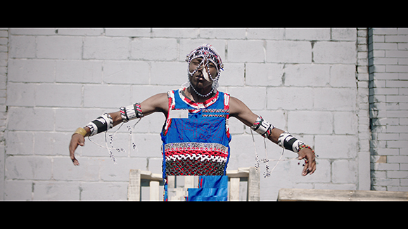 A person in blue and red traditional attire with beaded headpiece and armbands performs a dance.