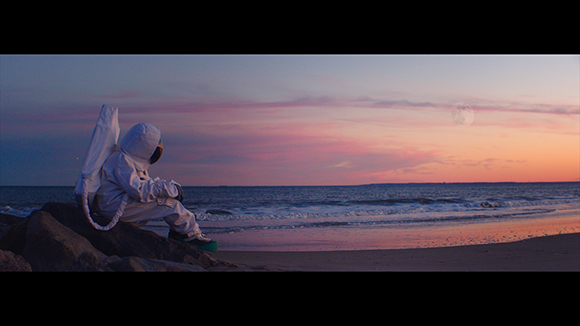 An astronaut in a white suit sits on dark rocks facing a sunset ocean with a large moon in the sky.