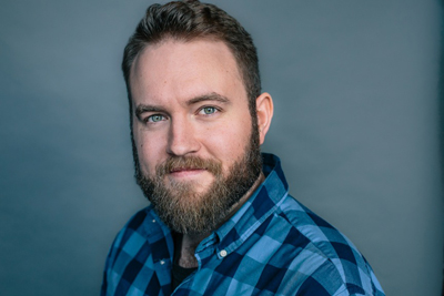 A man with light eyes and a brown beard wearing a blue checkered button-down shirt against a gray background.
