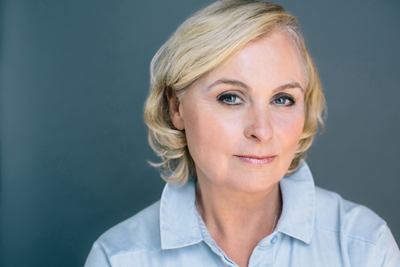 A blonde woman with blue eyes wearing a light blue collared shirt against a dark teal background.