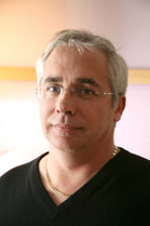 Close-up headshot of a middle-aged man with graying hair, wearing glasses and a black V-neck shirt with a gold chain.
