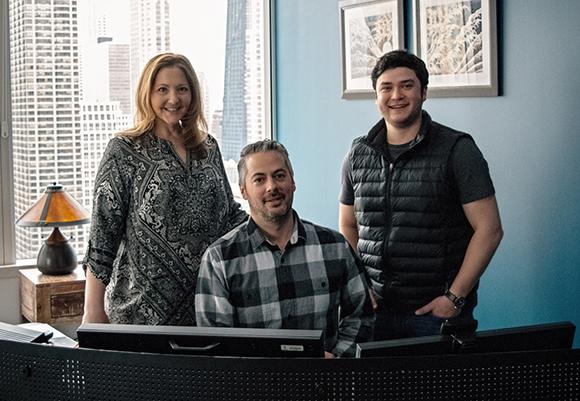 A woman in a patterned blouse, a man in a plaid shirt, and a man in a puffer vest pose in an office.