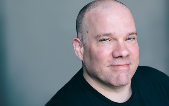 A close-up portrait of a bald man with light eyes and a slight smile against a gray background.