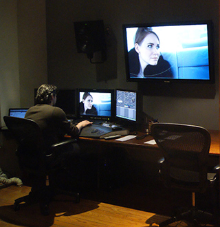 A person sits at a desk with three computer monitors, editing video in a dark room.