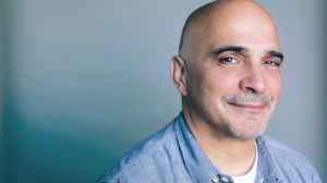 A bald man with a light beard and a blue button-down shirt smiles against a blue background.