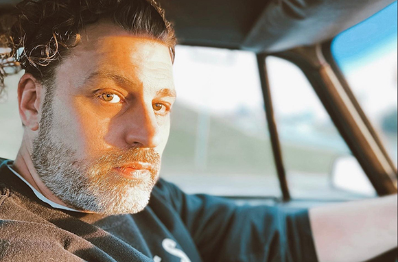 A man with curly dark hair and a salt-and-pepper beard looks forward while sitting in a vehicle.