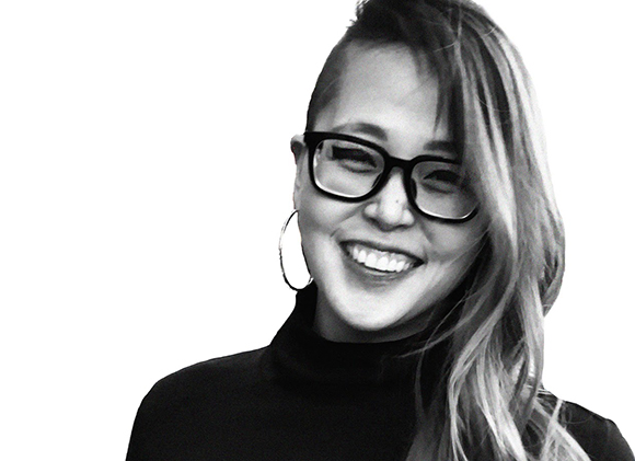 A smiling woman with long hair, glasses, and hoop earrings against a white background.