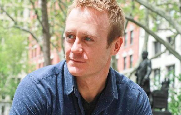 A man with reddish-blonde hair wearing a blue button-down shirt, looking off-camera in a park.