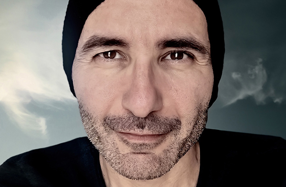 Close-up portrait of a man with dark eyes and stubble wearing a black beanie against a cloudy sky background.