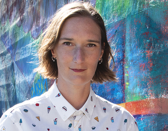 A person with shoulder-length brown hair wearing a white shirt with small colorful patterns against a blue mural.