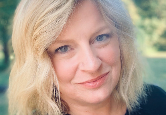 A blonde woman with blue eyes smiling at the camera in a soft-focus outdoor setting.