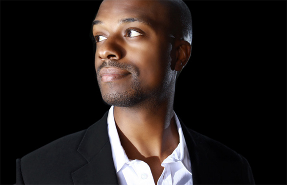 A man with short hair and a goatee looks off to the side against a black background.