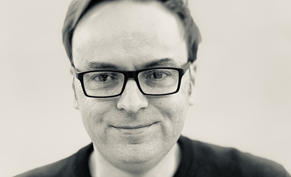 A close-up black and white portrait of a man with short hair wearing dark-rimmed glasses.