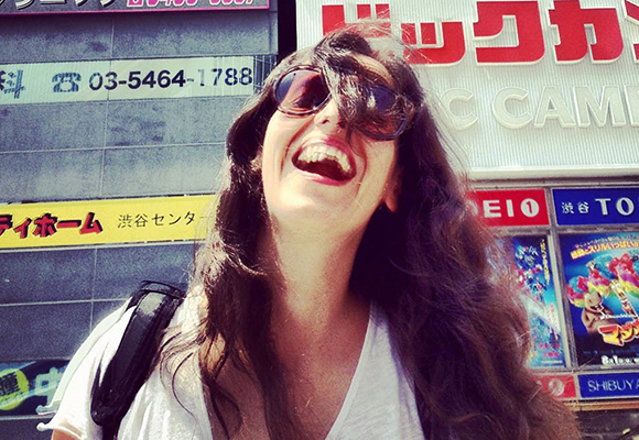 A woman with long dark hair and sunglasses laughs while looking upward in a city setting.