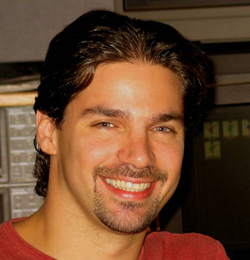 A smiling man with dark wavy hair and a goatee wearing a red shirt.