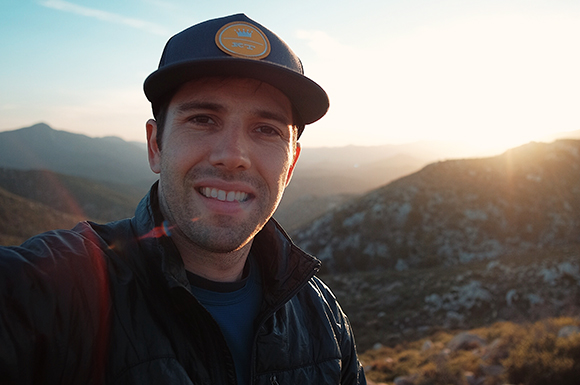 A man in a baseball cap and puffer jacket smiles during a sunset in a mountainous landscape.