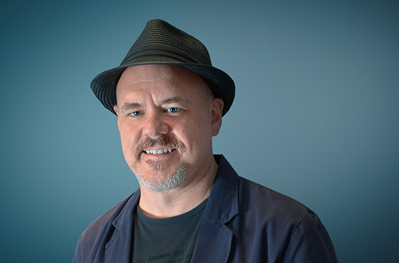 A smiling man with a goatee wearing a dark fedora and a blue jacket against a blue background.