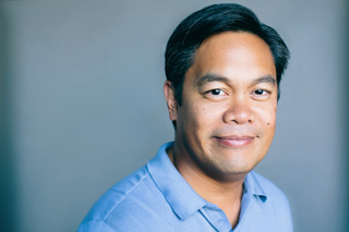 A man with short dark hair and a light blue polo shirt smiling against a gray background.