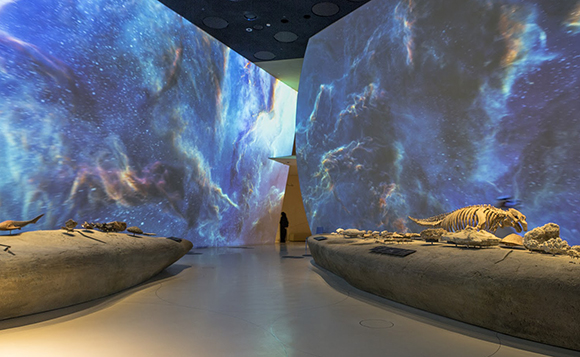 A person stands in a dark room surrounded by massive, curved screens displaying blue and purple nebula projections.