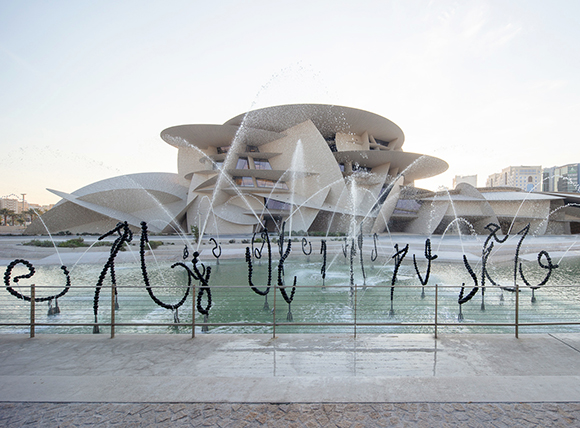 A large, white, multi-layered organic building behind a fountain featuring black calligraphic sculptures.