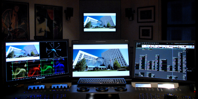 Three computer monitors on a desk displaying video editing software and building footage in a dark room.