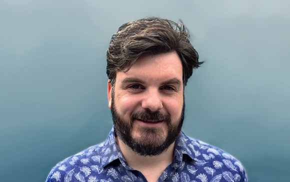 A man with dark hair and a beard wearing a blue patterned shirt, smiling against a solid blue background.