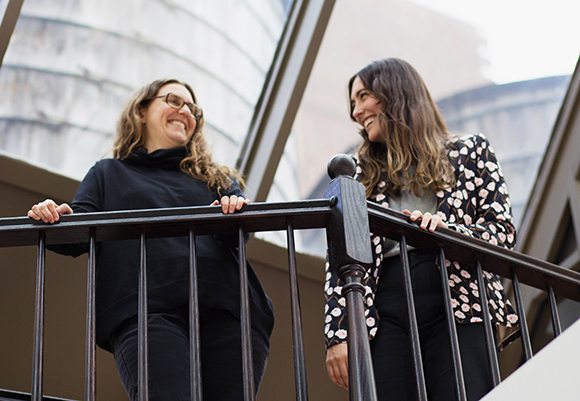 Two women laughing while standing on a balcony with a large building in the background.