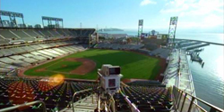 Aerial view of a baseball stadium with green grass, brown dirt, and seating next to a harbor with boats.