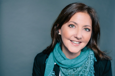 A smiling woman with brown hair and green eyes wearing a teal scarf against a gray background.
