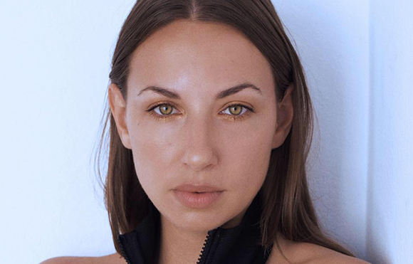 Close-up portrait of a woman with long brown hair and light-colored eyes looking directly at the camera.