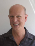 A smiling bald man wearing a dark patterned button-down shirt against a plain light background.