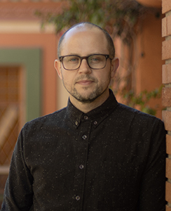 A man with glasses and a short beard wearing a black patterned button-down shirt.