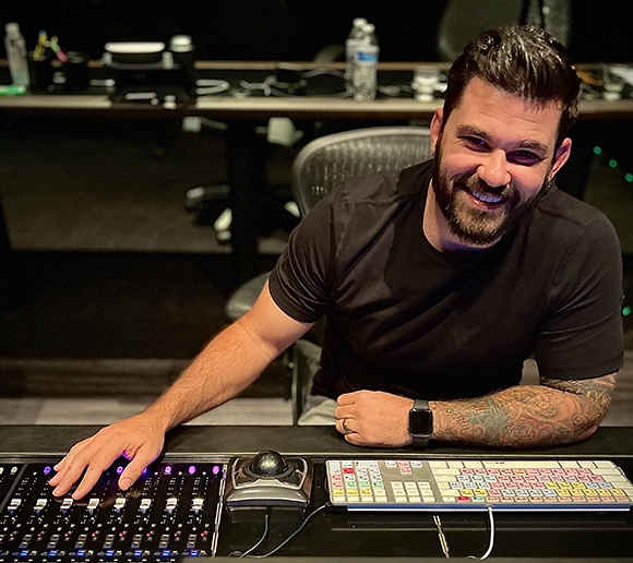 A smiling man with dark hair and a beard sits at a professional audio mixing desk in a studio.