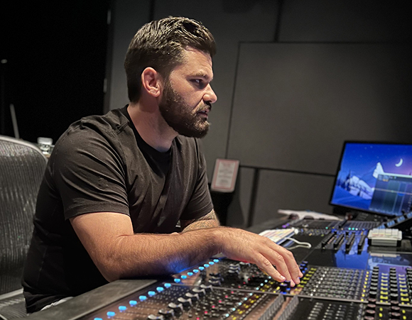 A man with a beard sits at a large audio mixing console in a dark studio setting.