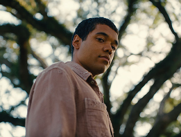 A young man in a tan button-down shirt looks toward the camera with trees in the background.