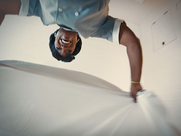 A smiling person in a light blue uniform leans over a large white sheet from a low angle.