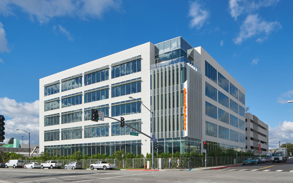 A large, modern white office building with many windows at a city intersection under a blue sky.