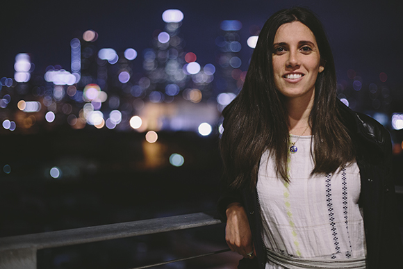 A smiling woman with long dark hair wearing a white patterned shirt and black jacket against a night city bokeh.