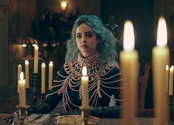A woman with blue hair wearing an elaborate pink beaded collar sits at a table with lit candles.