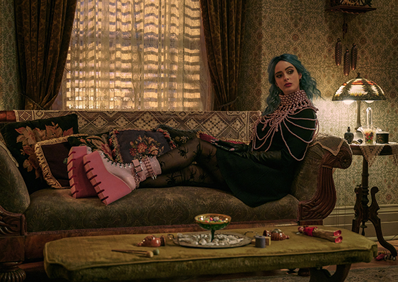 A woman with blue hair in a black dress and pink boots reclines on a patterned sofa in a dimly lit room.