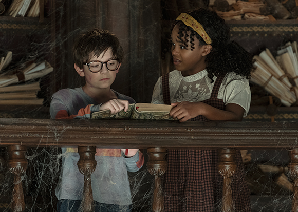 A boy with glasses and a girl in a checkered dress look at an open book in a cobweb-filled attic.