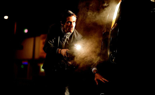 A man in a dark jacket holds a bright flashlight at night next to a vehicle with smoke or steam rising.