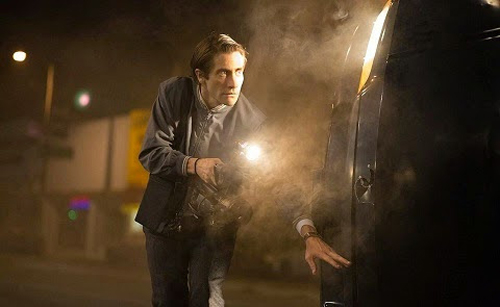 A man leans forward, shining a bright flashlight into the open door of a dark van at night.