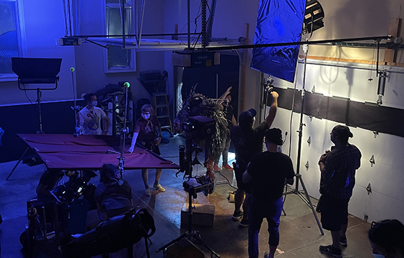 Film crew members surround a large creature puppet under studio lights in a dark studio.